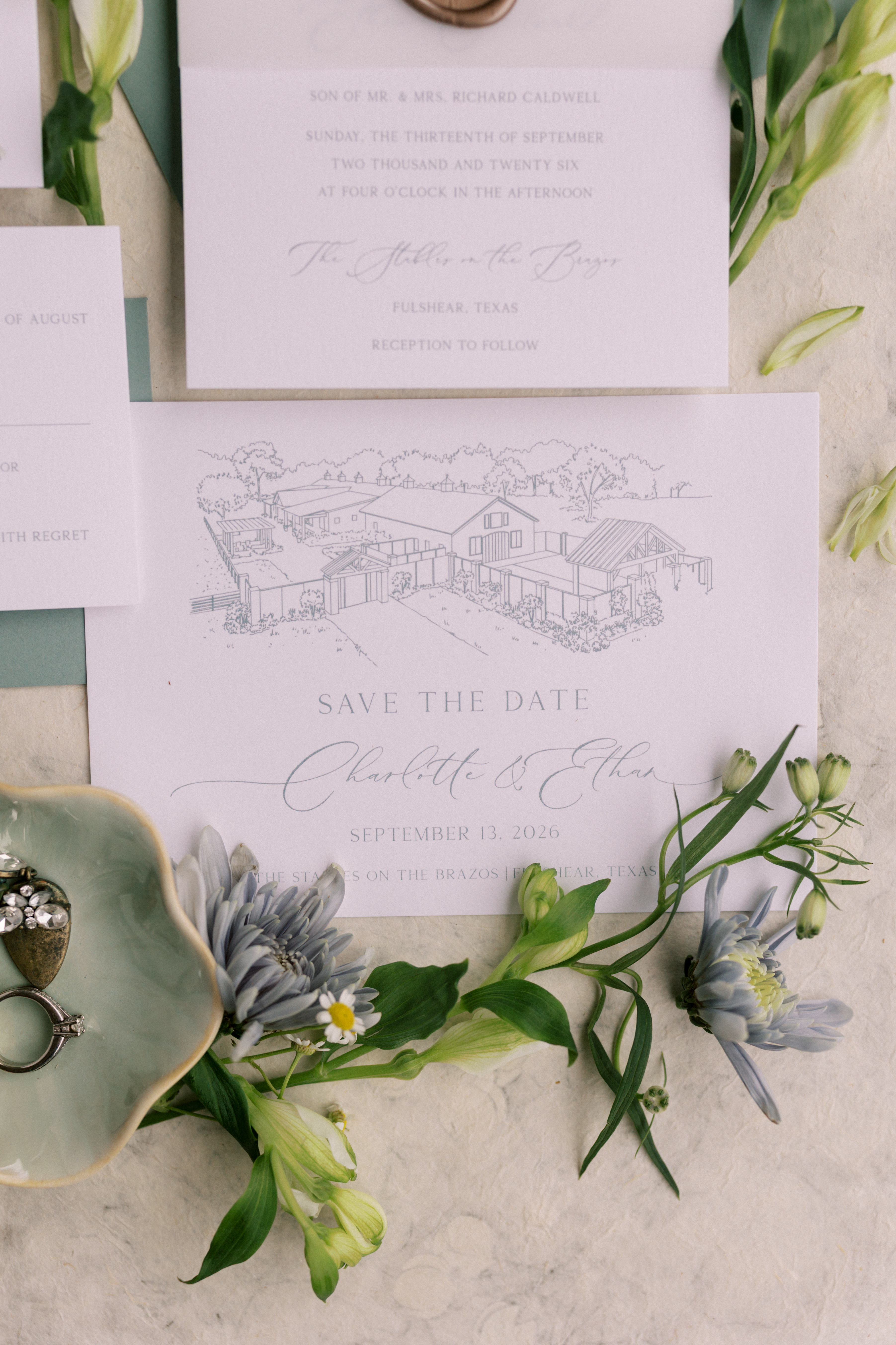 Save the date card with a detailed line drawing of a barn venue, styled with pale green envelopes and blue-grey flowers