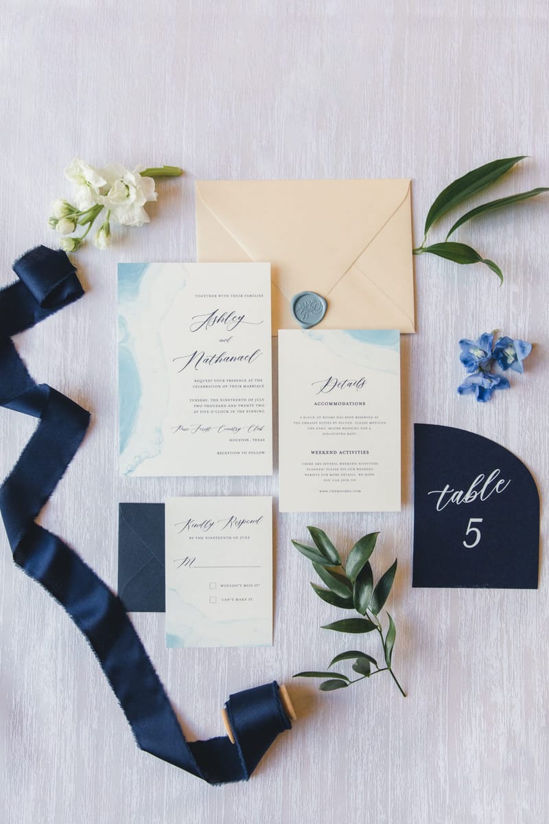 Light blue watercolor wedding invitation suite with a gold envelope and navy ribbon, including an RSVP card, details card, and table number, styled with white and blue flowers