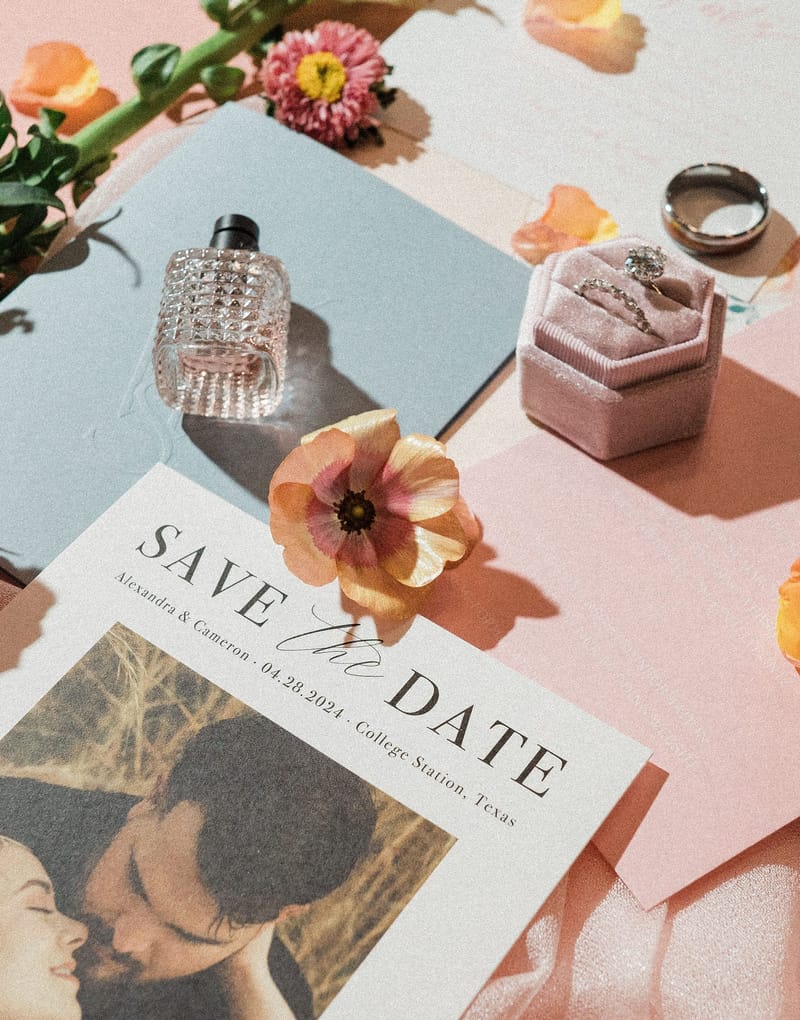 Custom save the date card with a couple's engagement photo, styled with a blue envelope, a pink ring box, and a perfume bottle on a pink surface with flowers