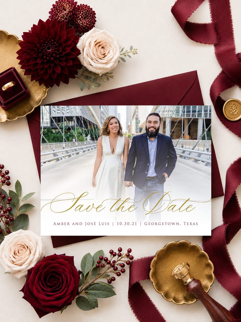 Burgundy save the date card with an engagement photo of a couple, styled with burgundy ribbon, a gold wax seal stamp, and red and white flowers